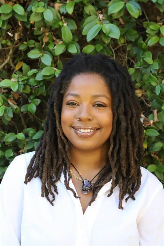 Kalayah, a Basic Needs CalFresh Coordinator, smiling at the camera in front of a leafy background